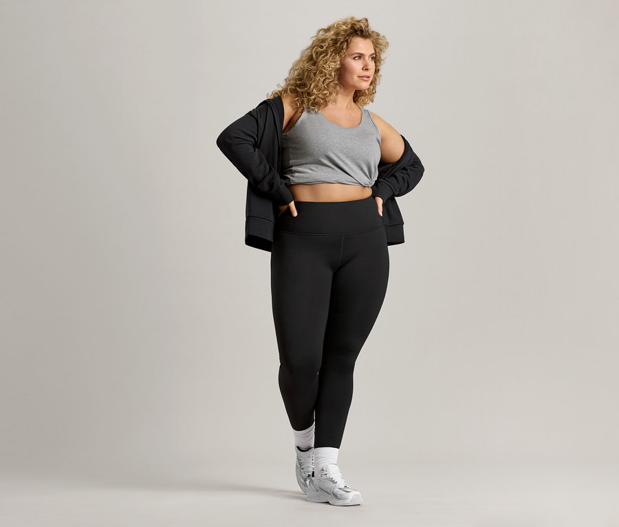 Žena s kudrnatými vlasy má na sobě šedé tílko, černou bundu a černé legíny.