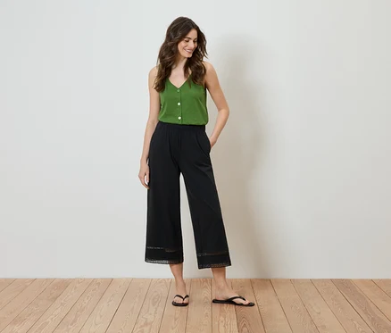 Žena stojí s rukama v kapsách, má na sobě zelený top a černé culottes kalhoty s krajkovým detailem na lemu.