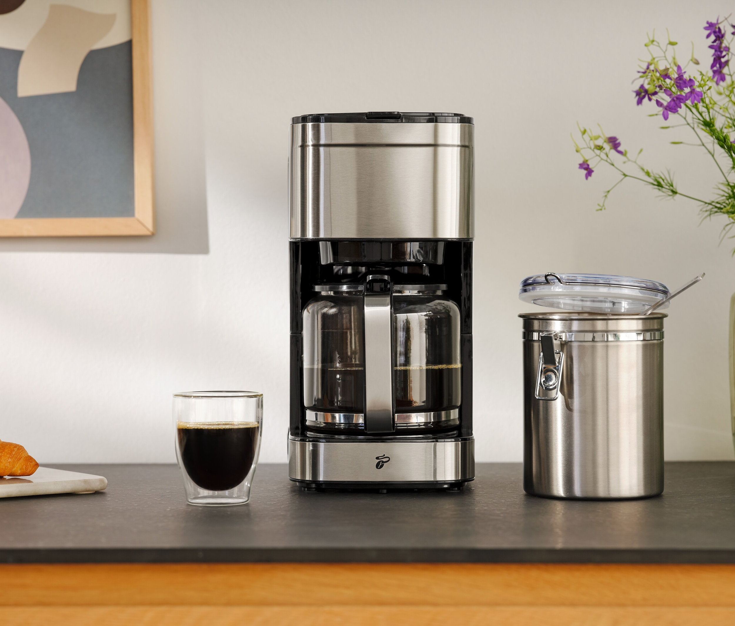 Kávovar vedle sklenice kávy, croissantu a skladovací nádoby.