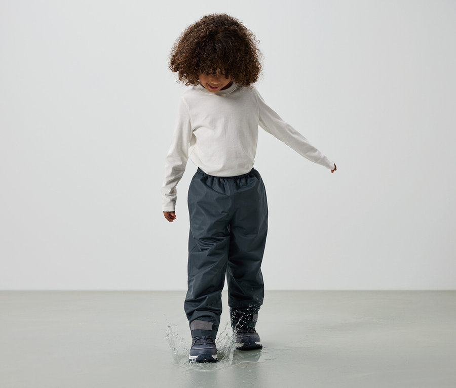 Dítě s kudrnatými vlasy, oblečené v tričku s dlouhým rukávem, šedých dešťových kalhotách a botách, skáče do kaluže.