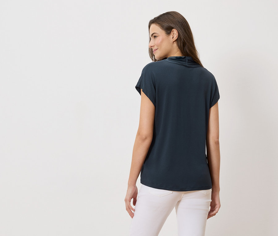 Pohled zezadu na ženu s dlouhými hnědými vlasy v tmavě modré halence a bílých džínách.