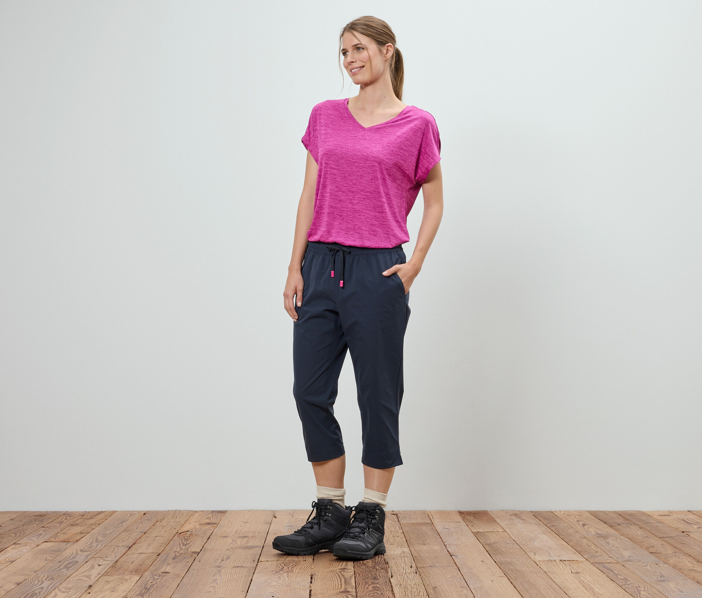 Žena stojí s rukama v kapsách před bílou stěnou. Má na sobě růžové tričko, modré tříčtvrteční kalhoty a černé trekové boty.
