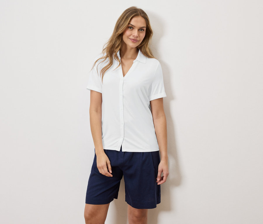 Žena stojí před zdí a má na sobě bílou košili a modré šortky.