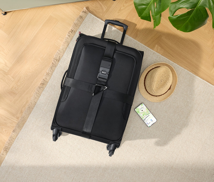 Černý kufr na kolečkách s kloboukem a smartphonem na béžovém koberci.
