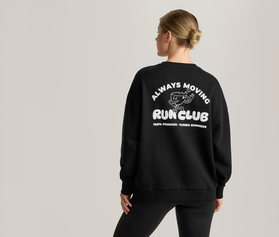 Žena zezadu má na sobě černou mikinu s bílou grafikou. Grafika obsahuje kreslenou postavičku šálku kávy a text 'ALWAYS MOVING RUN CLUB' a '100% PASSION - TCHIBO RUNNERS'.