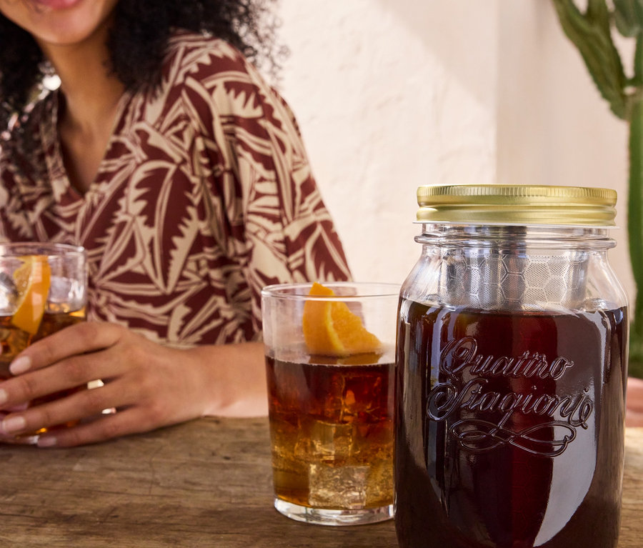 Žena drží sklenici s nápojem vedle sklenice pro přípravu cold brew »BrewJar« a žerzejových šatů s potiskem.