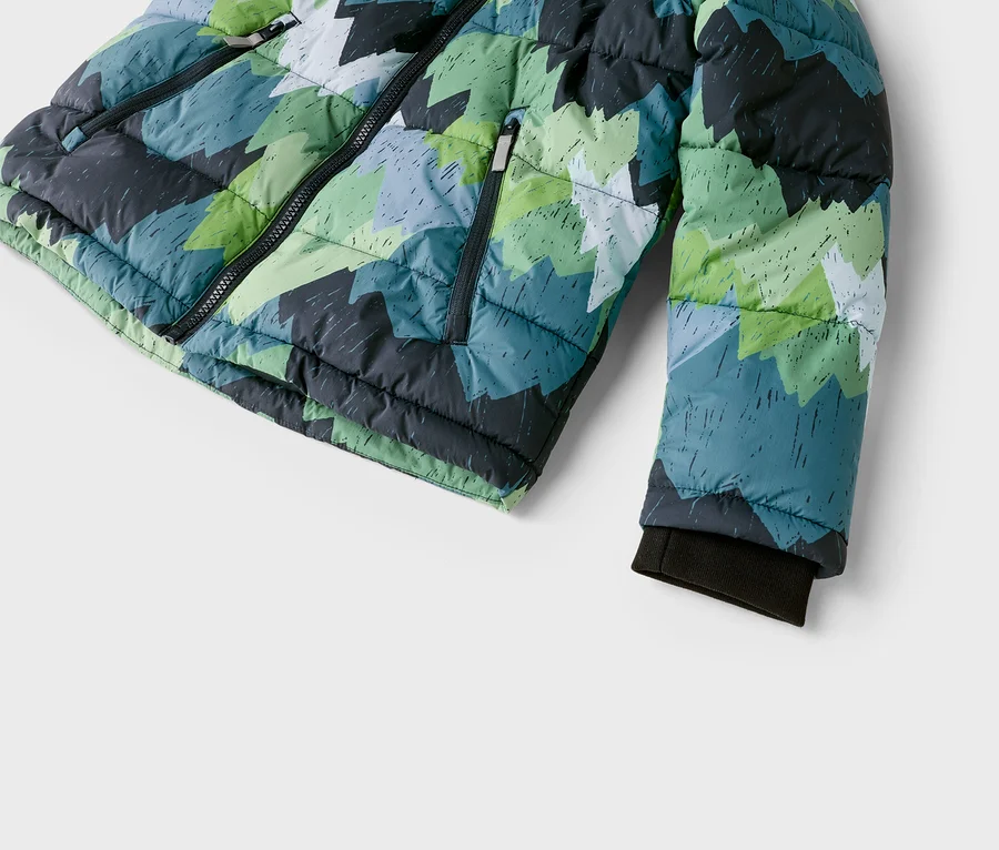 Detail prošívané bundy s geometrickým vzorem zelených, modrých, šedých a bílých zubatých tvarů. Bunda má černý přední zip, kapsu na zip a tmavou žebrovanou manžetu na rukávu.