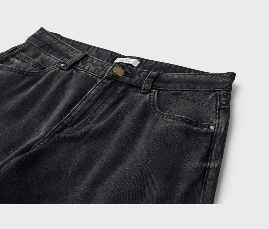 Detail horní části černých džínů z opraného denimu. Džíny mají pas s poutky na opasek, zapínání na zlatý knoflík a přední kapsy, včetně malé kapsičky na mince. Bílý štítek s nápisem „MY STYLE“ je viditelný u knoflíku.