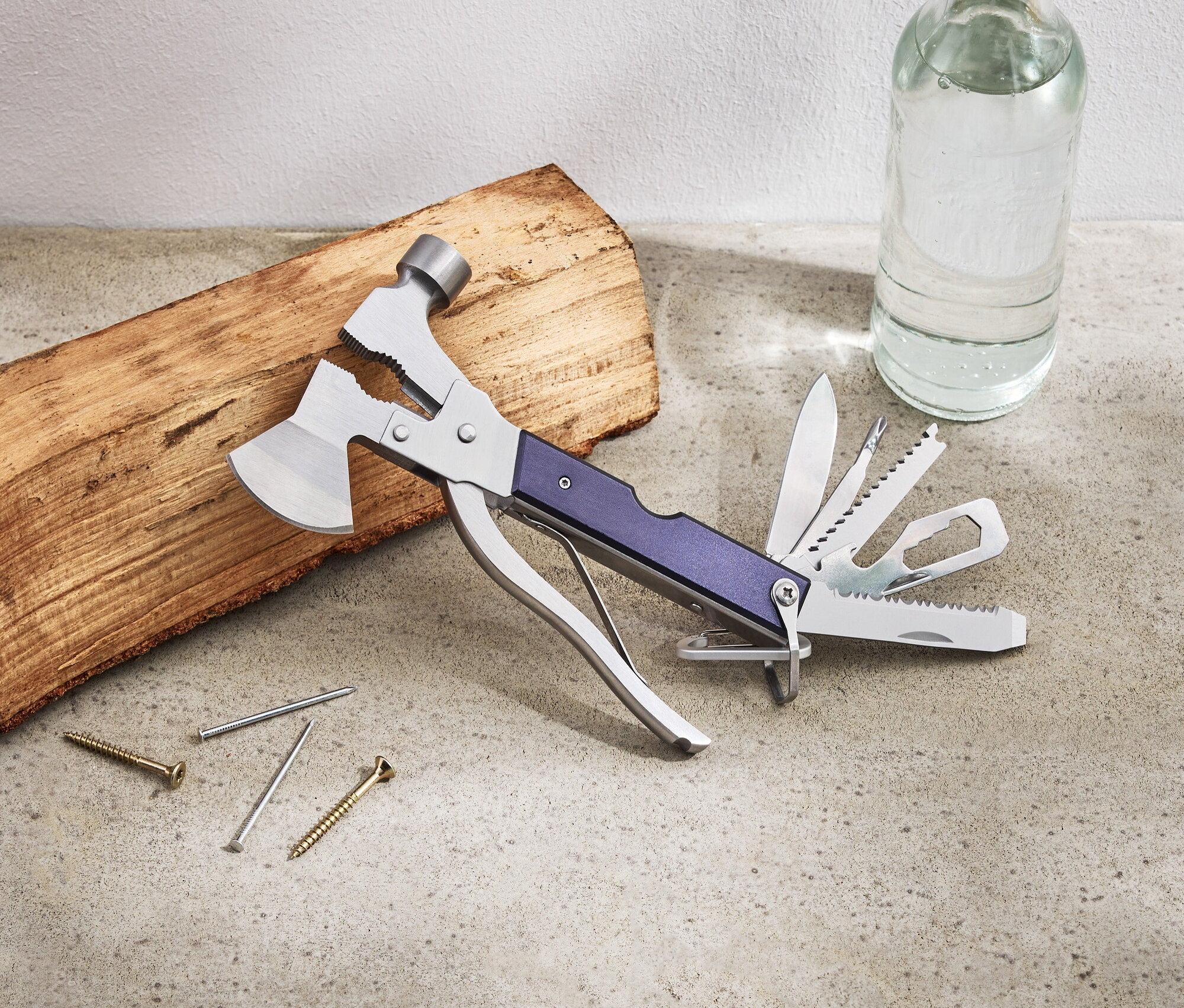 Na stole leží multifunkční nástroj, kus dřeva, hřebíky, šrouby a láhev s vodou.