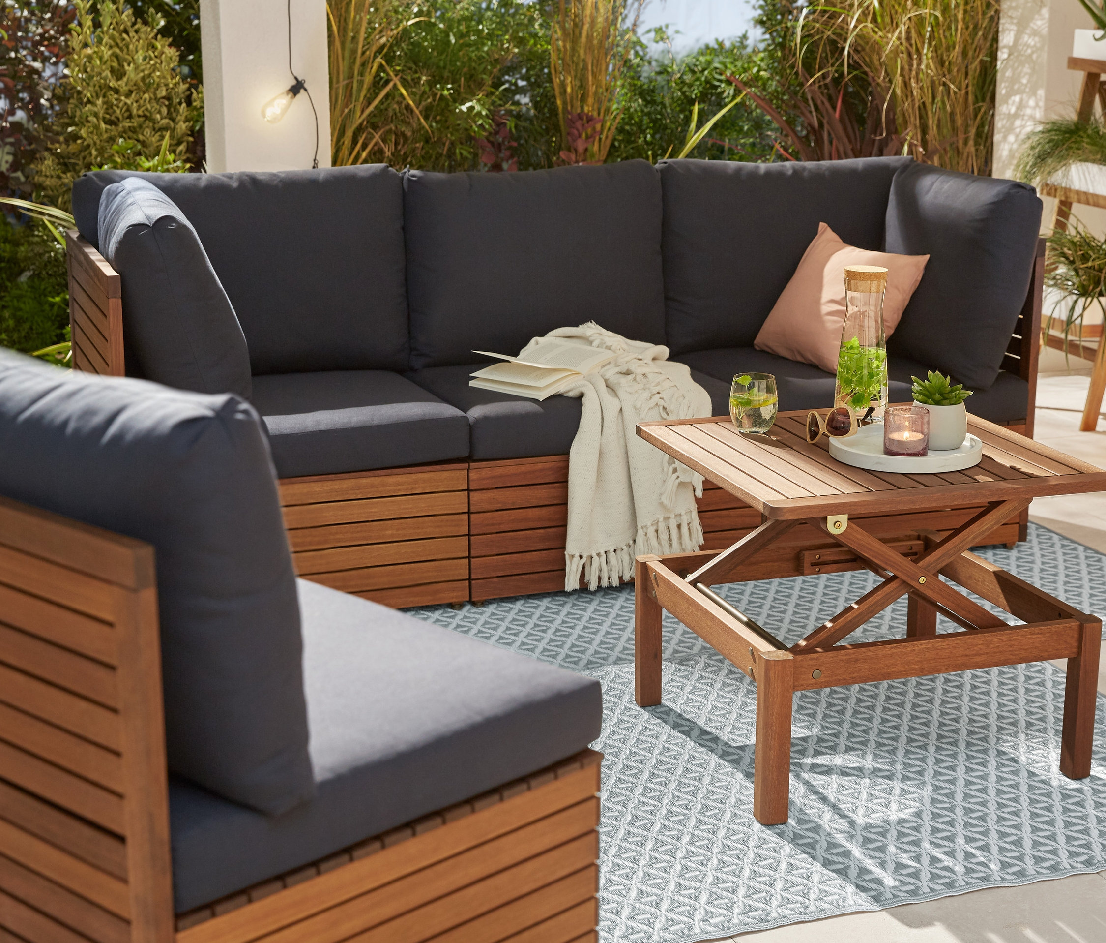 Rohová sedací souprava s křeslem a zahradním stolkem na venkovním koberci. V pozadí je žebřík na rostliny a lucerna s LED žárovkami.