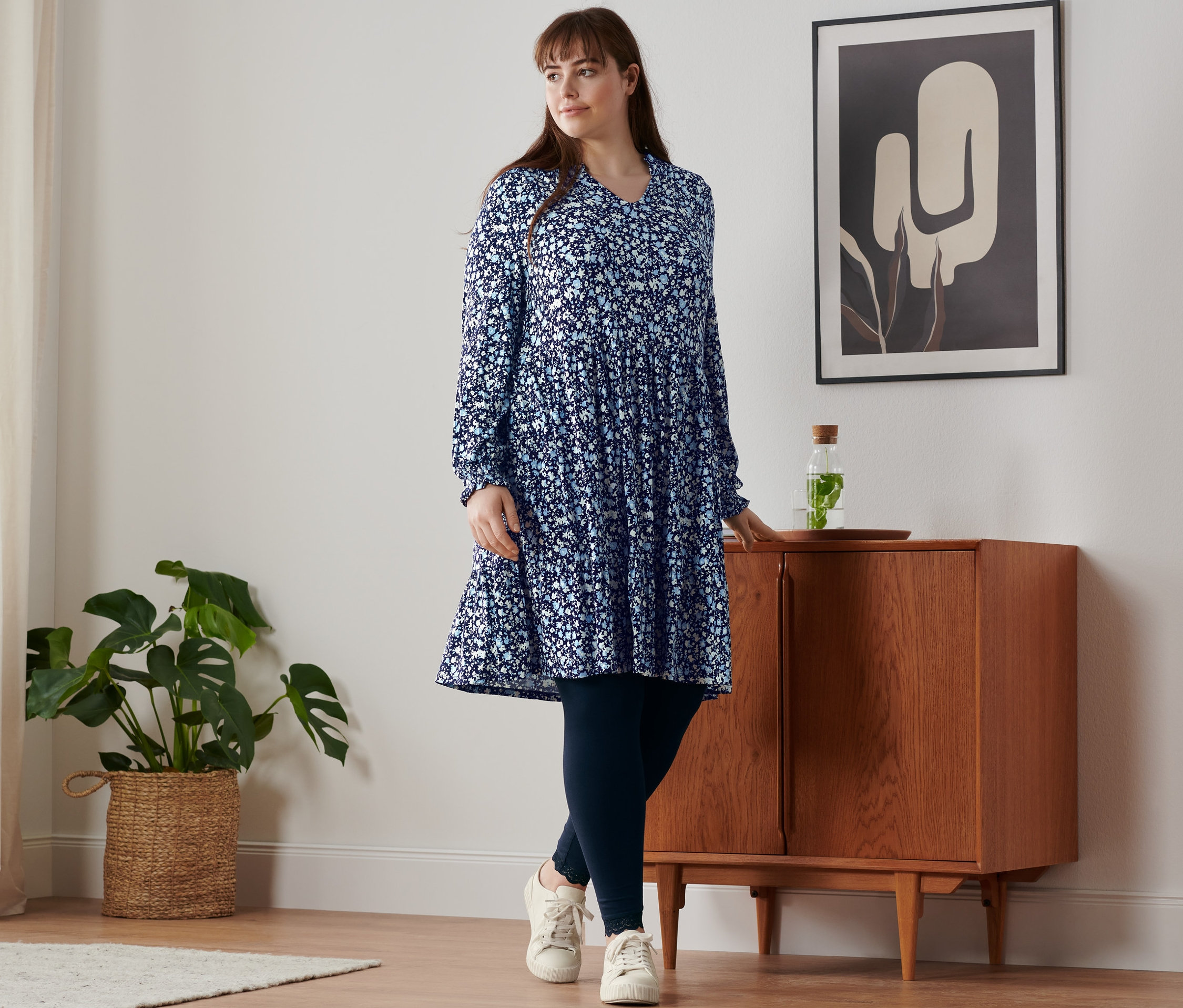 Žena stojí u skříňky a má na sobě květované žerzejové šaty, legíny s krajkovým lemem a kožené boty sneaker.