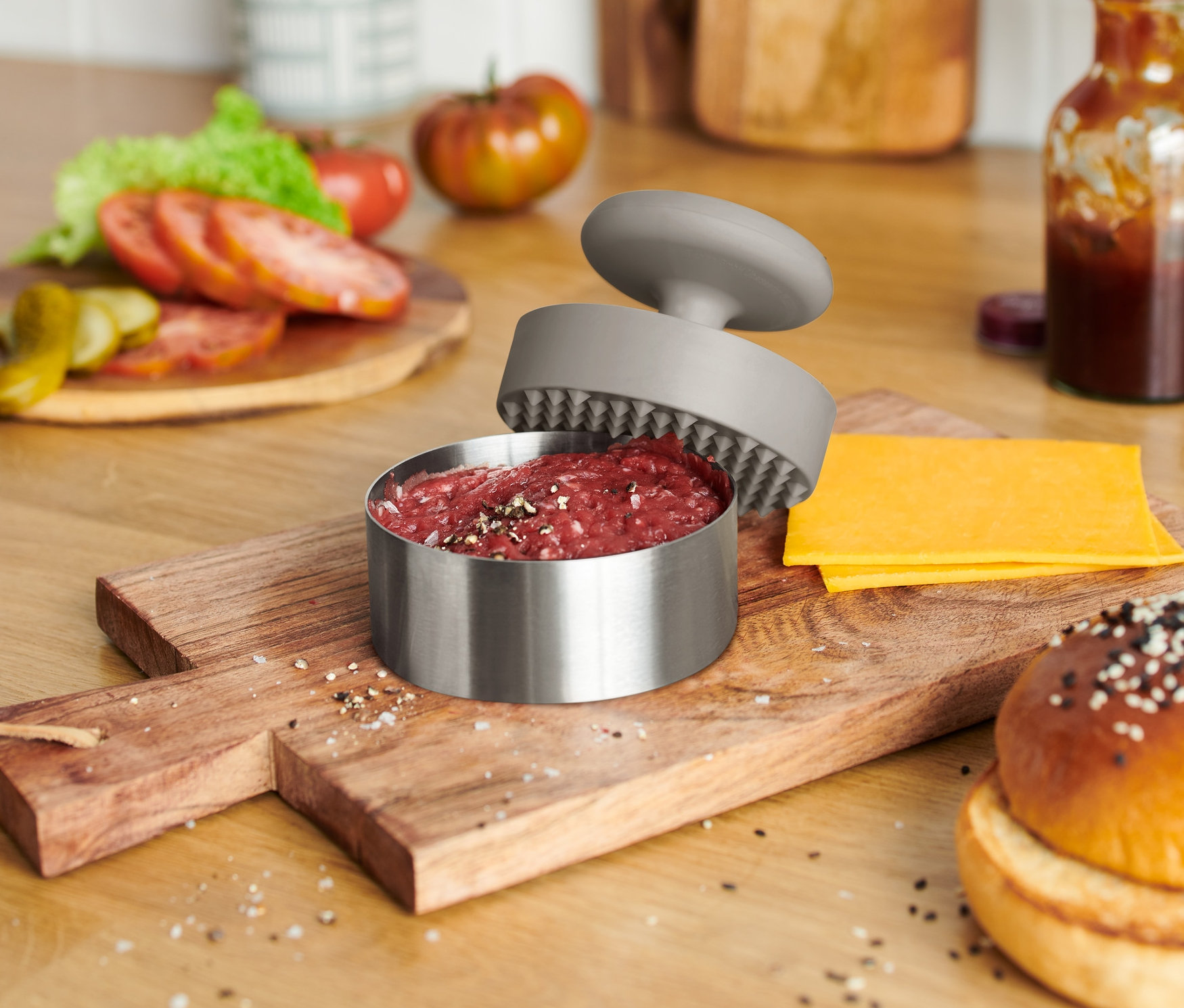 Na dřevěném prkénku je hamburgerová houska, sýr a otevřený lis na hamburgery s masem.