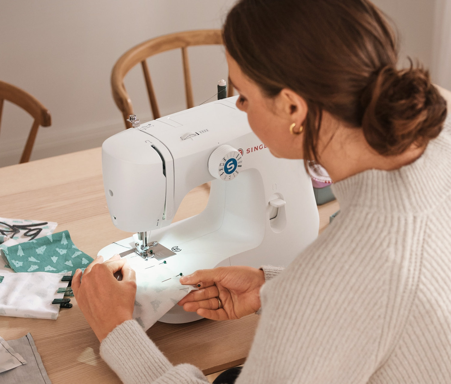 Žena šije látku na šicím stroji SINGER vedle sady šicích nití, sady na šití »Látkový adventní kalendář« a úložných boxů.
