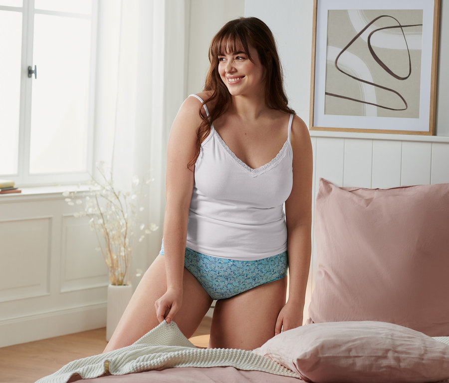 Žena klečí na posteli a drží deku. Má na sobě bílé tílko a kalhotky.