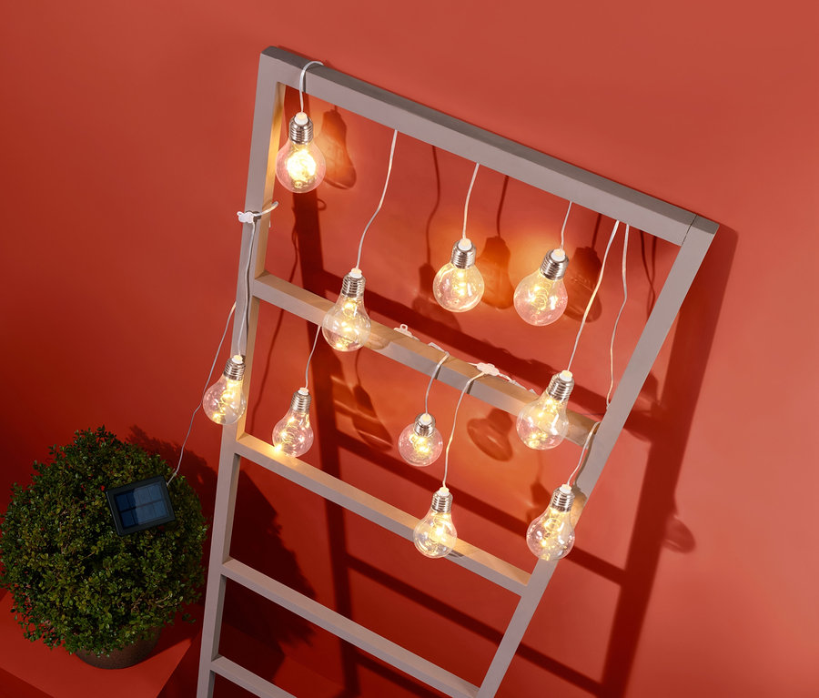 Solární světelný řetěz je zavěšený na šedém rámu, vedle malého keře na oranžovém pozadí.
