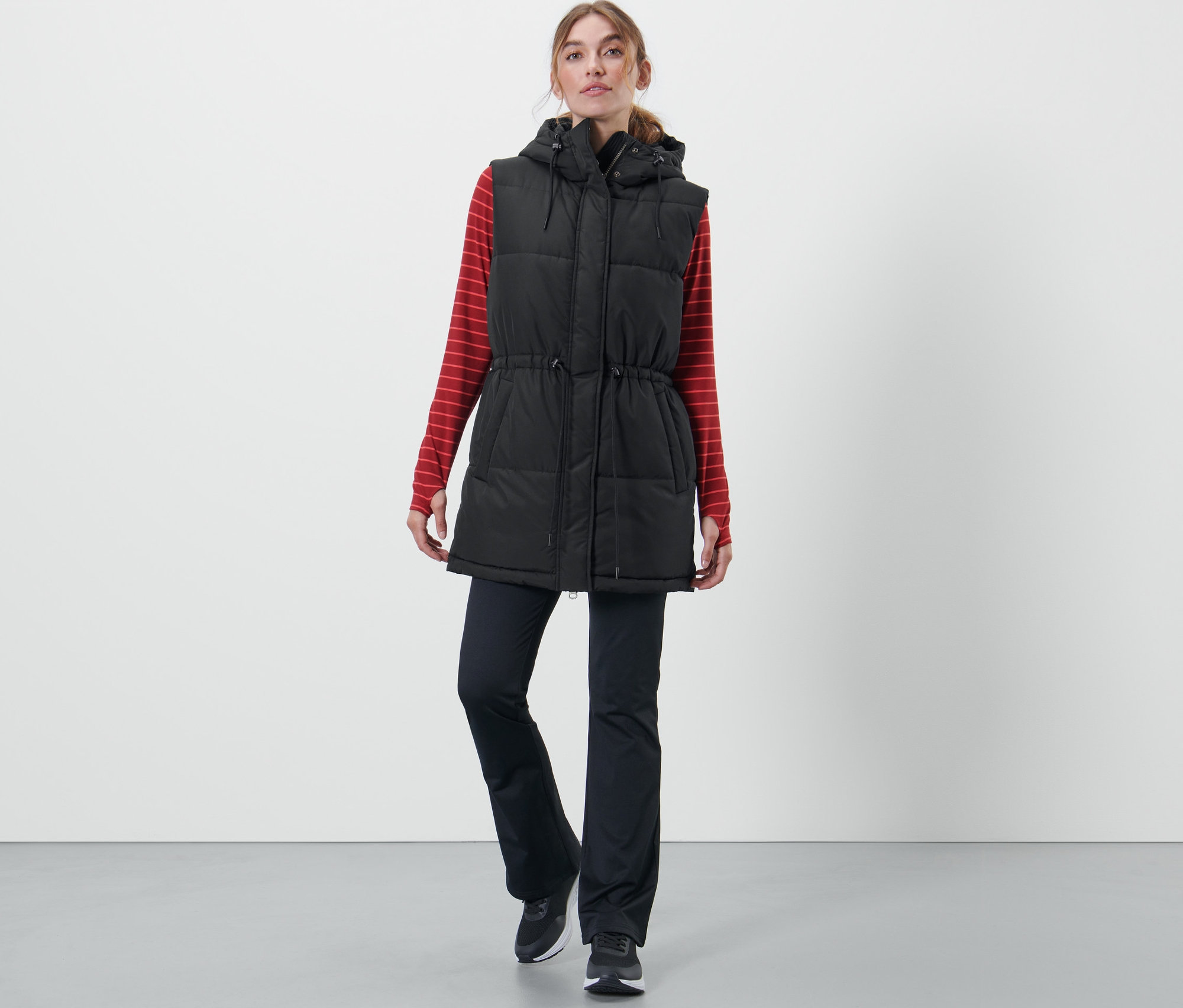 Žena má na sobě prošívanou vestu s kapucí, červené tričko s dlouhým rukávem a černé kalhoty.
