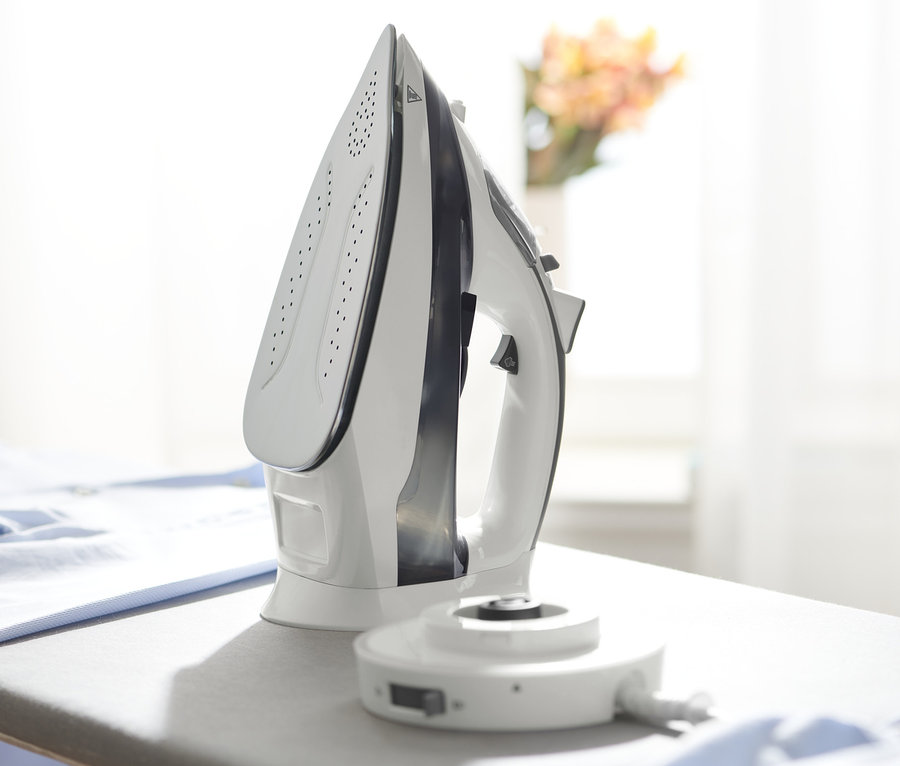 Bílá bezdrátová napařovací žehlička stojí na šedém žehlicím prkně vedle hromady modrého oblečení.