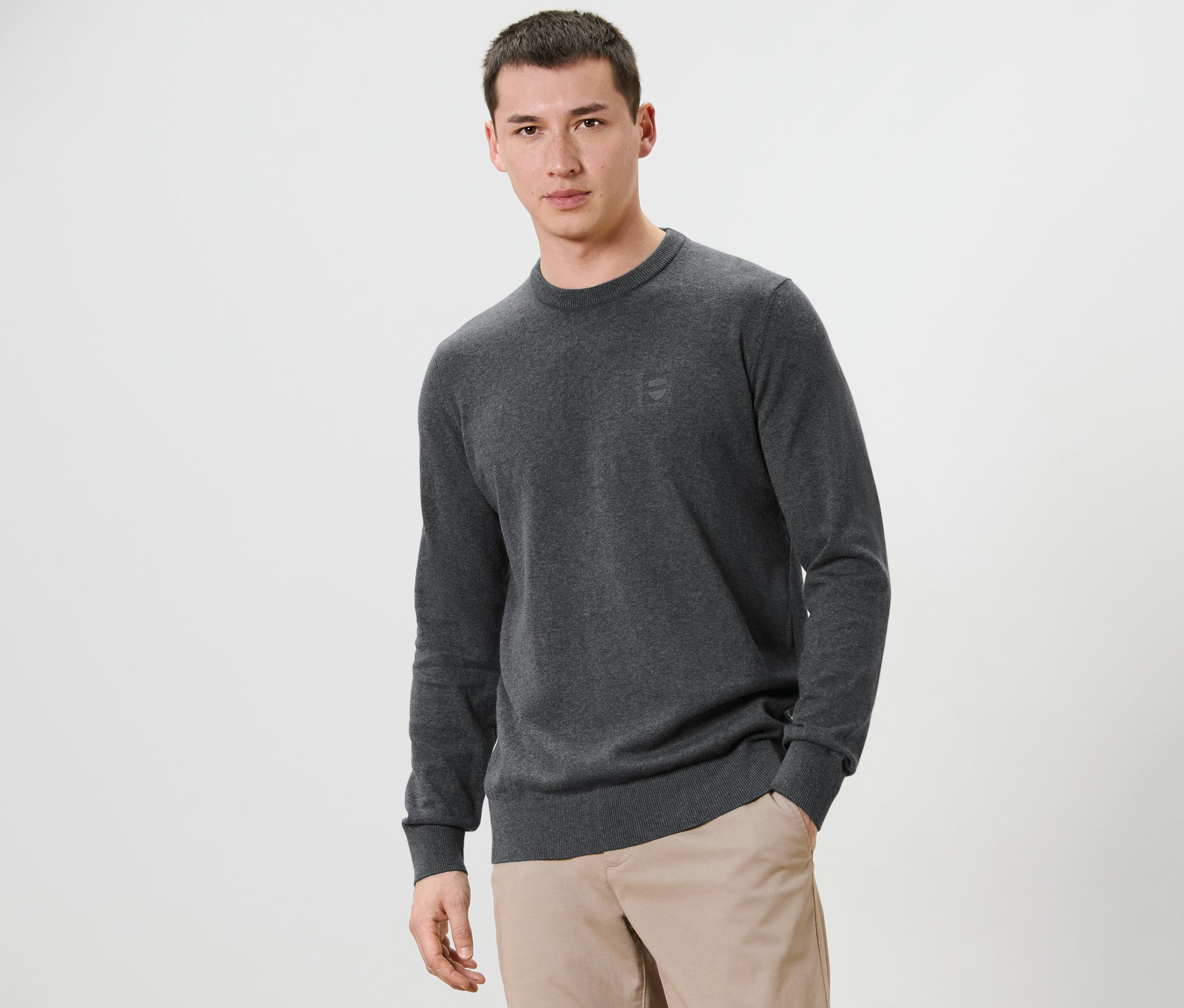 Muž pózuje na bílém pozadí a má na sobě šedý svetr s kulatým výstřihem a béžové kalhoty.
