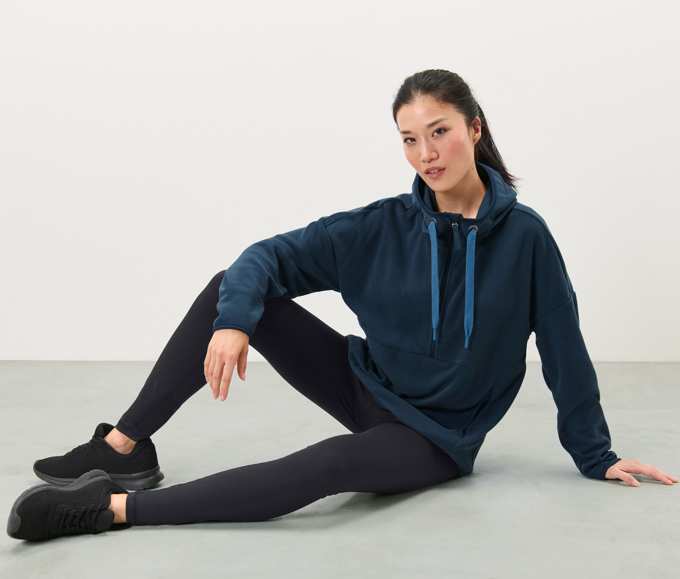 Žena sedí na zemi a má na sobě fleecový svetr s kapucí, černé legíny a tenisky.