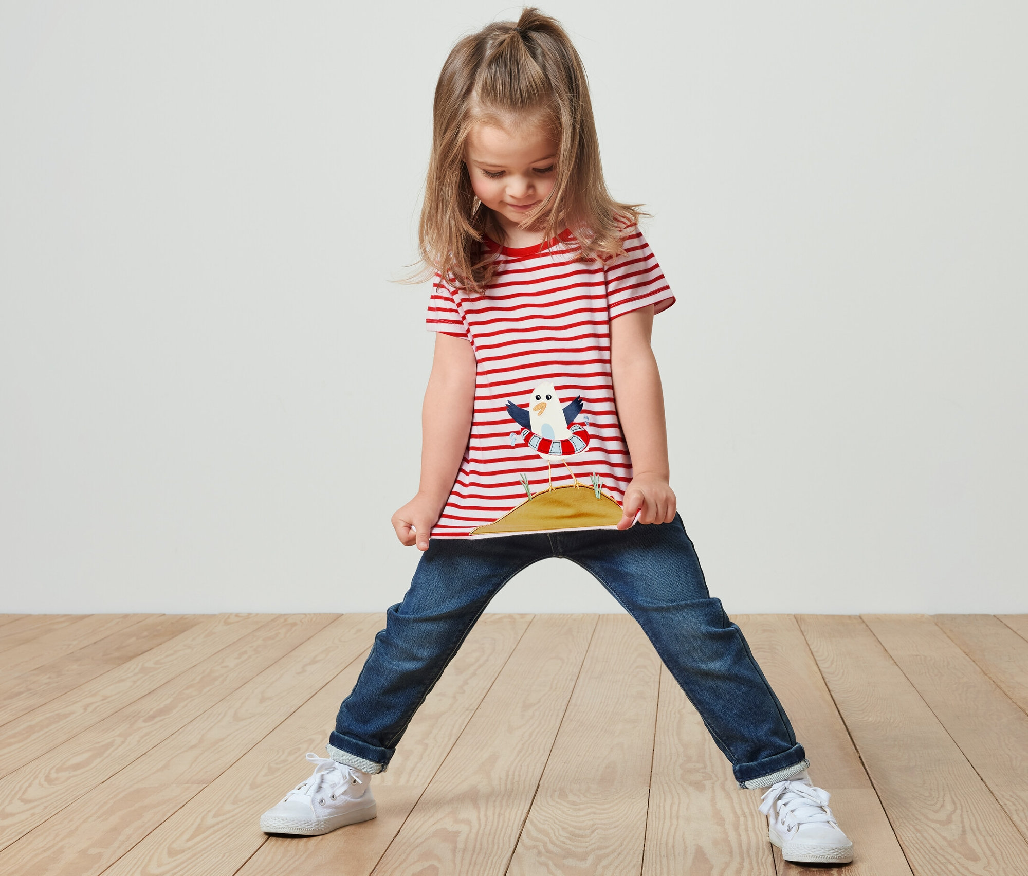 Dívka stojí a drží si okraj trička s červenými pruhy a aplikací.