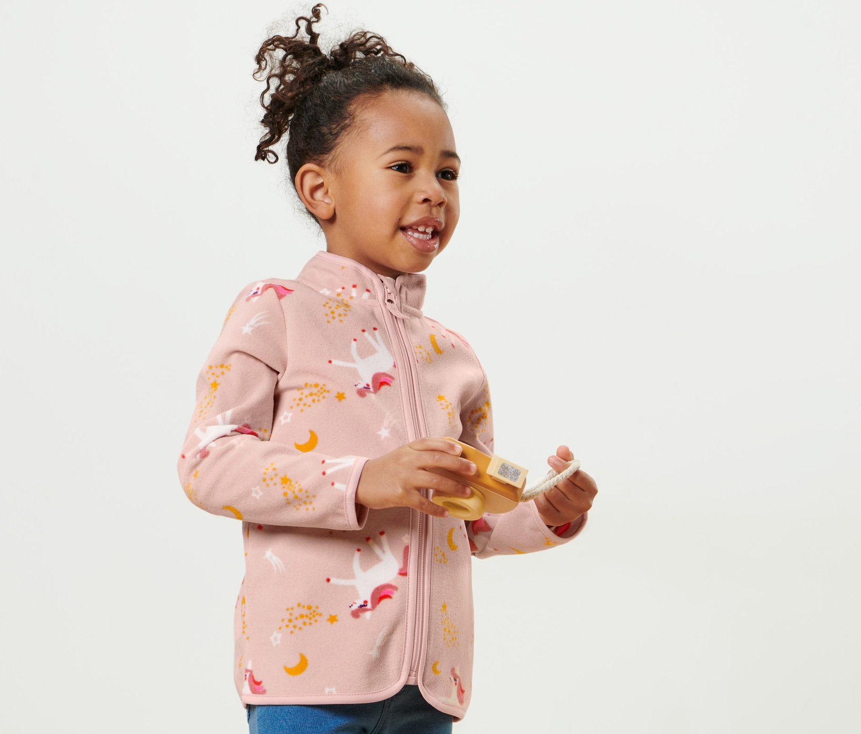 Usmívající se holčička má na sobě dětskou fleecovou bundu s jednorožcem a hraje si s hračkou.