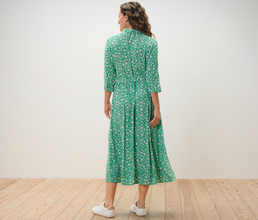 Žena stojí zády k fotoaparátu a má na sobě zelené košilové šaty s potiskem.