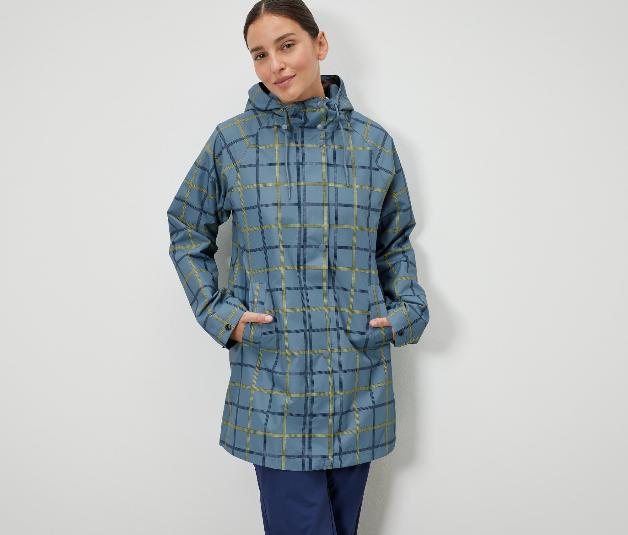 Žena stojí a má ruce v kapsách modré bundy do deště s kostkovaným vzorem.
