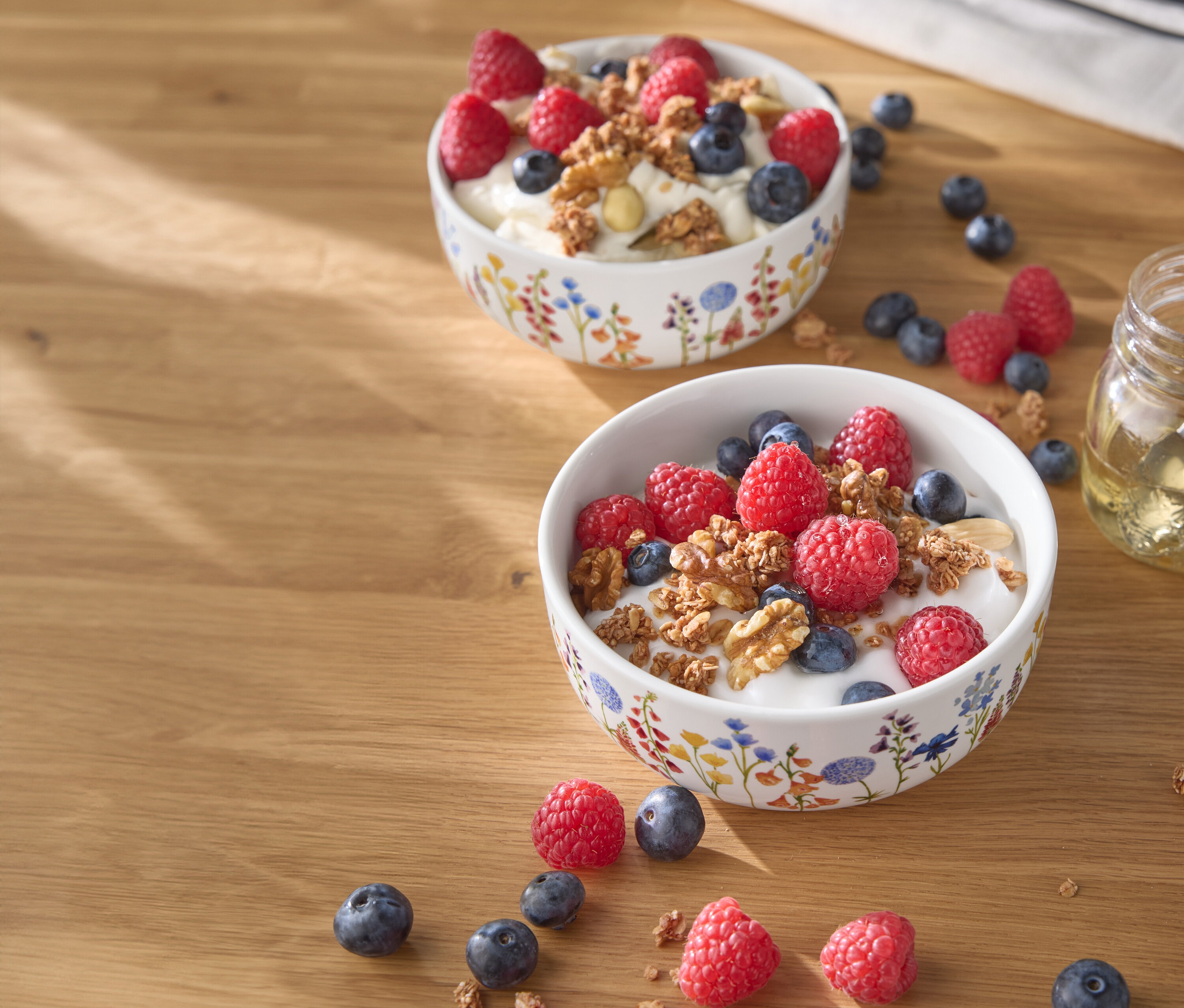 Dvě misky jogurtu s malinami, borůvkami a ořechy na dřevěném stole.