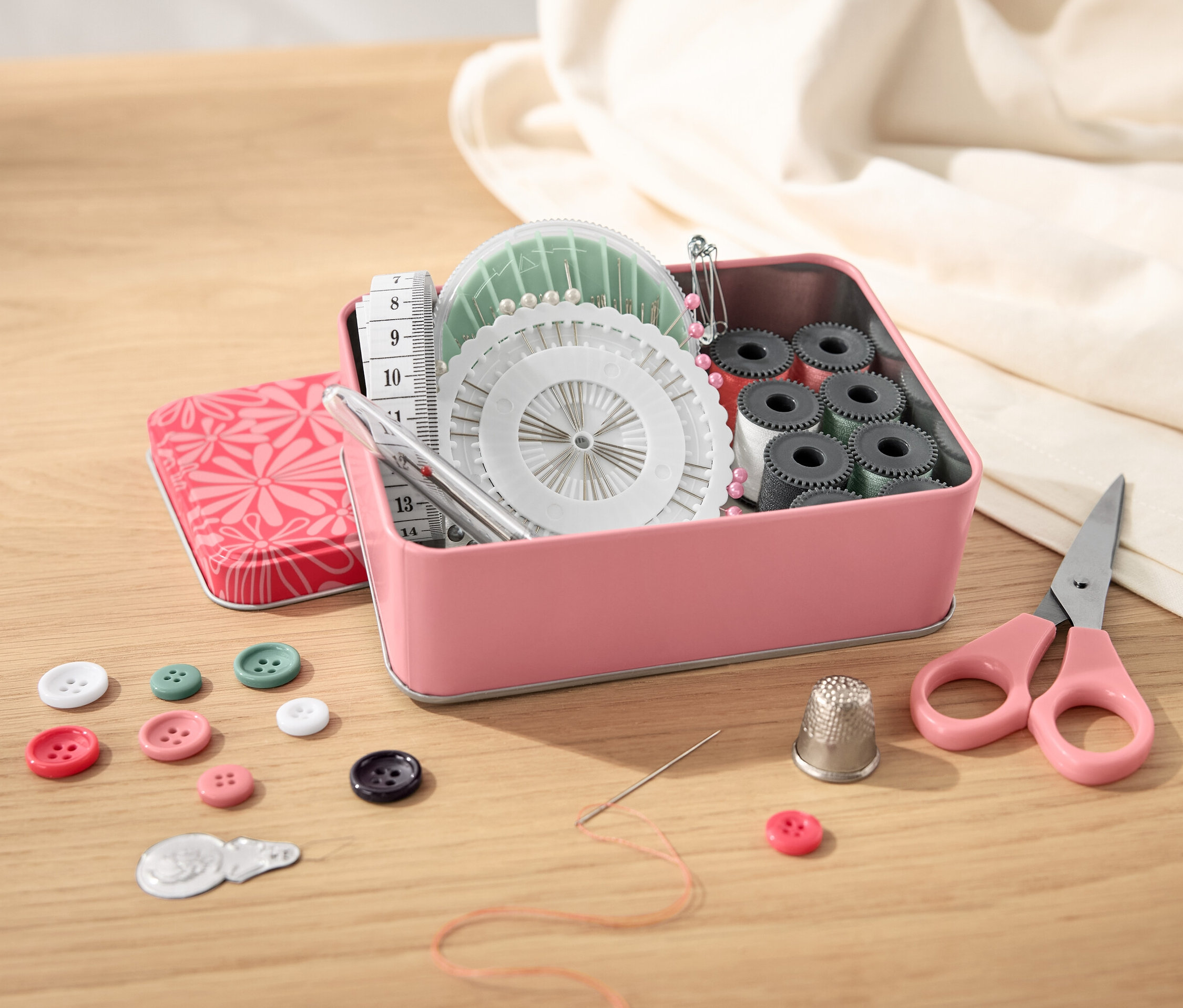 Otevřená kovová krabička s šicí sadou, knoflíky, nůžkami a krejčovským metrem na dřevěném stole.