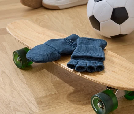 Modré prstové rukavice leží na dřevěném skateboardu s zelenými kolečky. V pozadí je fotbalový míč a boty.