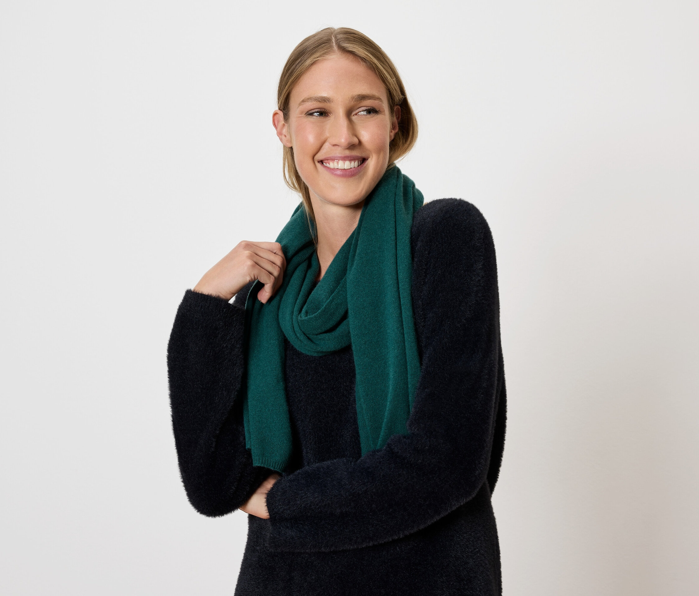 Žena s blond vlasy a úsměvem pózuje na světlém pozadí s tmavě zelenou šálou a svetrem.