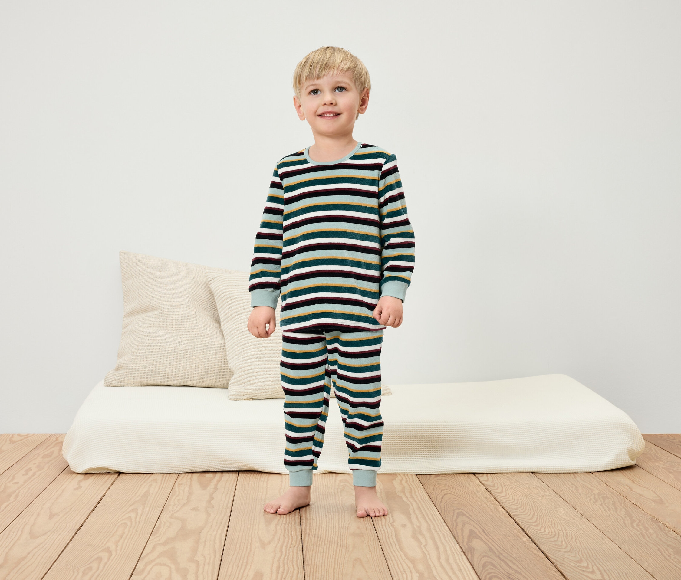 Chlapec v pruhovaném pyžamu stojí bosý na dřevěné podlaze před bílou zdí.