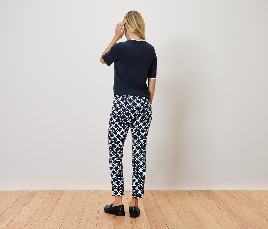 Žena stojí zády k fotoaparátu a má na sobě tmavě modrý top, vzorované kalhoty a černé boty.