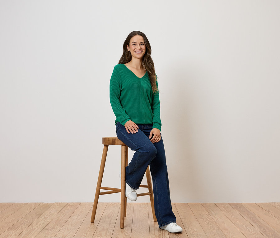 Žena sedí na dřevěné stoličce, má na sobě zelený svetr a džíny.