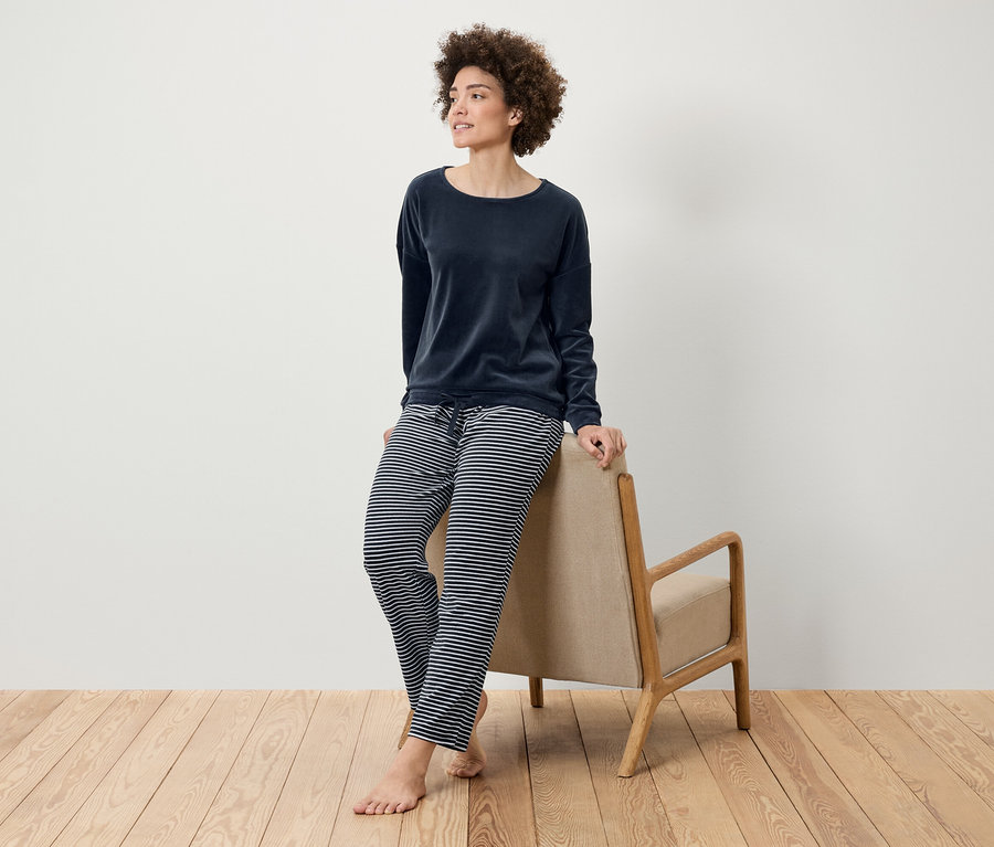 Žena s afro účesem pózuje v tmavě modrém sametovém topu a pruhovaných kalhotách, opírajíc se o béžové křeslo na dřevěné podlaze.