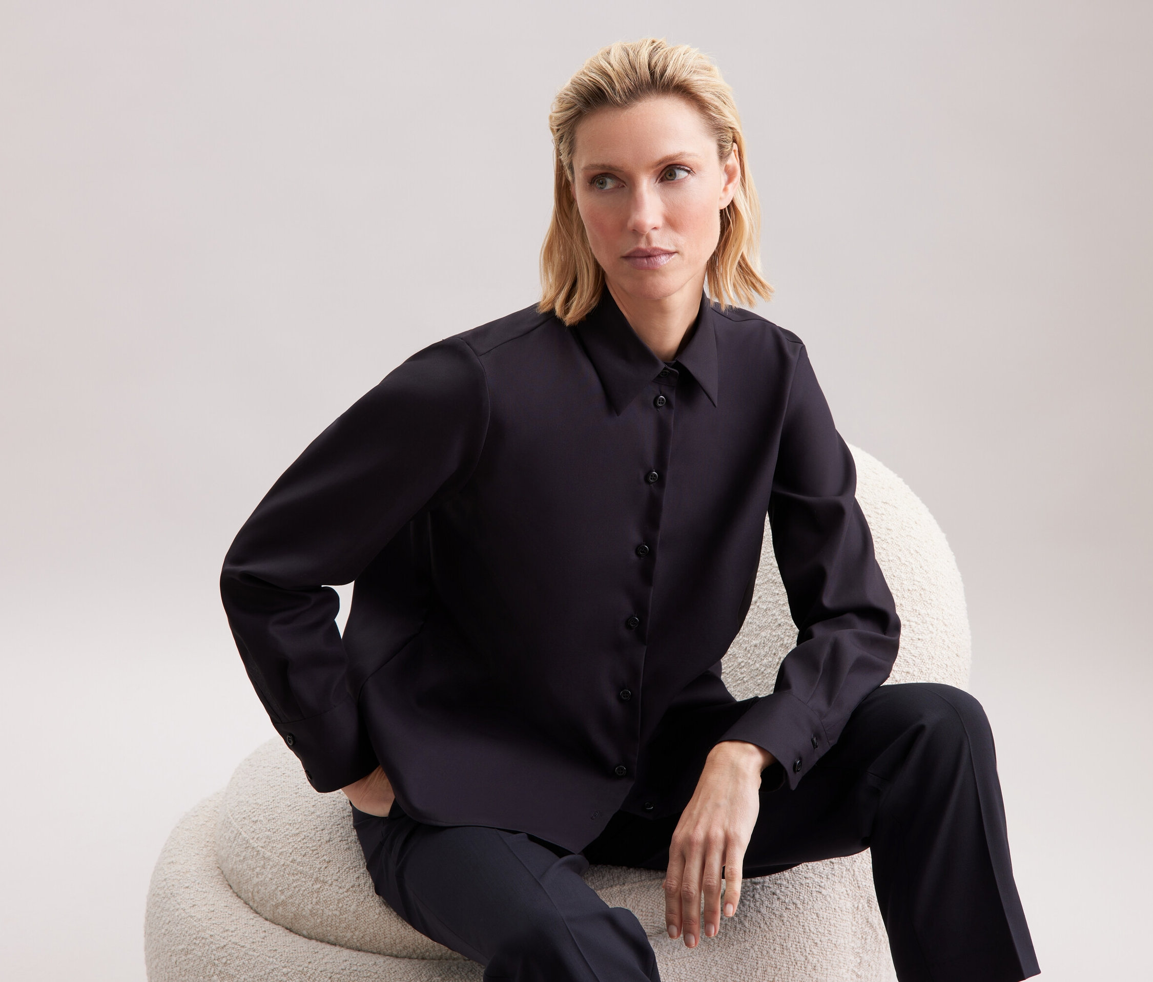 Blondýna sedí na béžovém křesle, má na sobě černou košili a kalhoty.