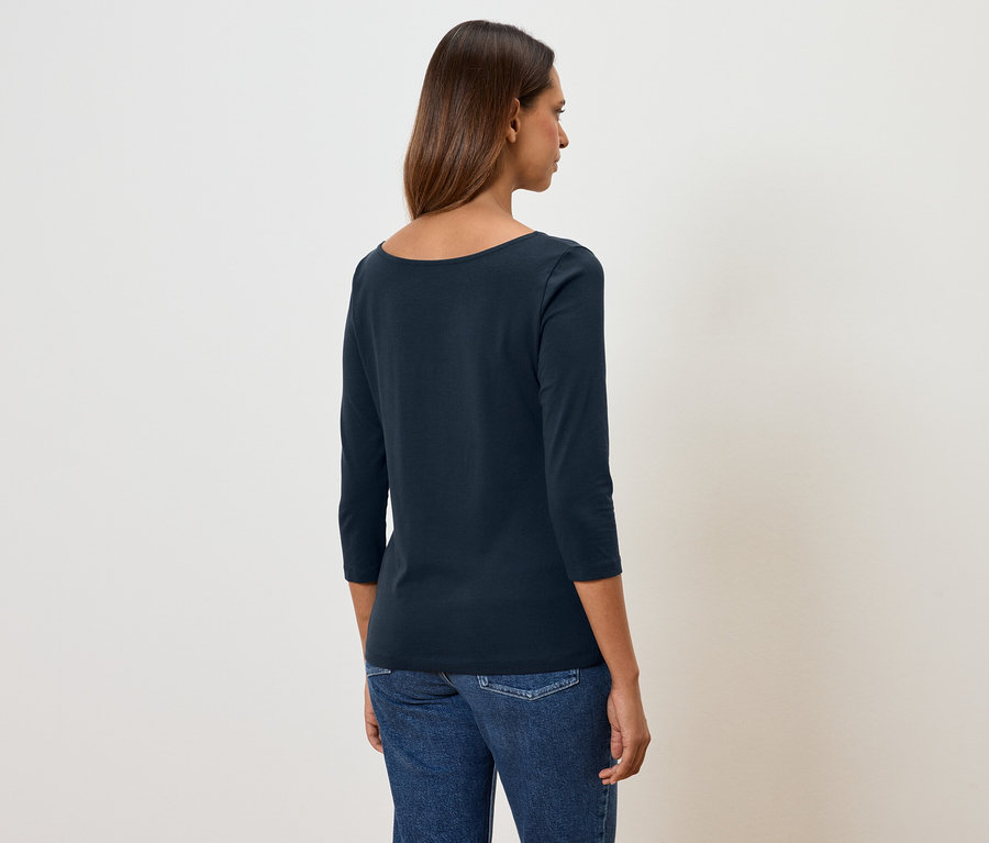 Žena zády k fotoaparátu má na sobě modré tričko s tříčtvrtečním rukávem a džíny.