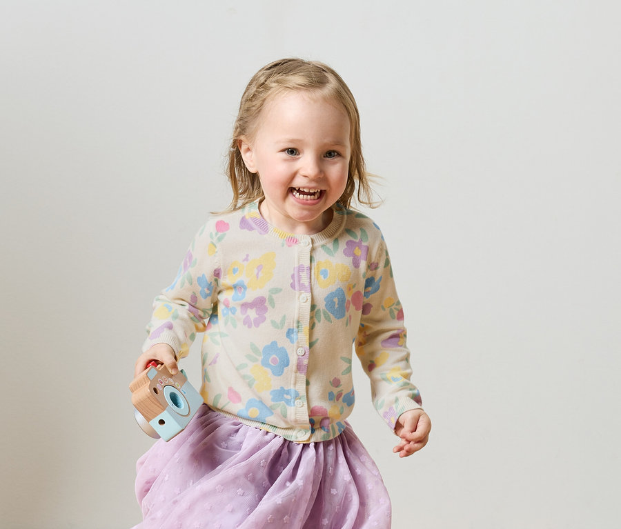 Usmívající se blondýnka se zapletenými copy v květovaném kardiganu a fialové sukni drží hračkářský fotoaparát.