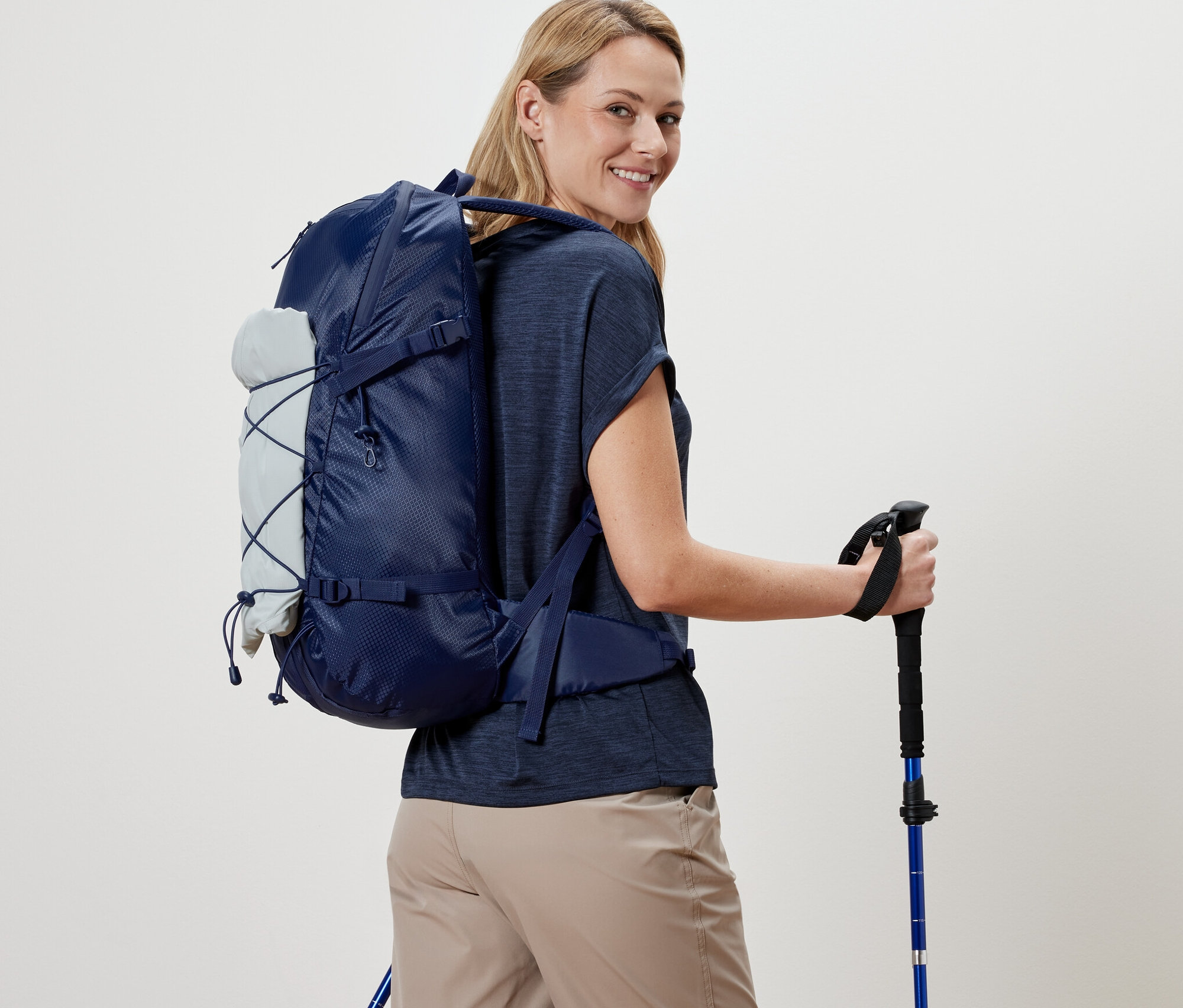 Žena s batohem a trekingovými holemi se ohlíží přes rameno.