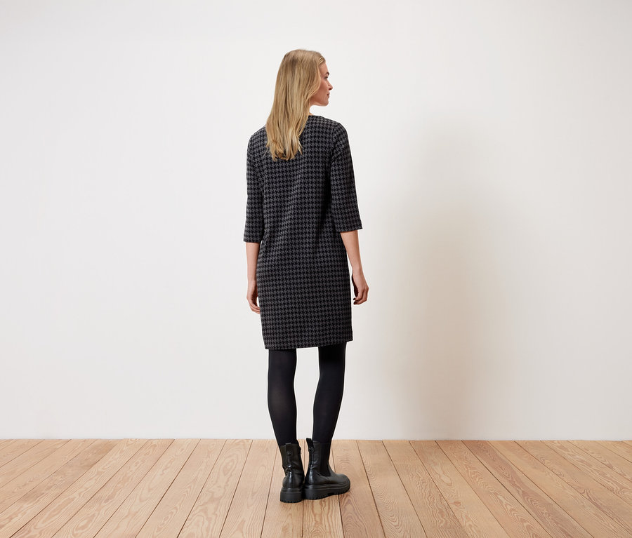 Žena stojí zády k fotoaparátu a má na sobě šaty s kohoutí stopou, černé legíny a černé boty.