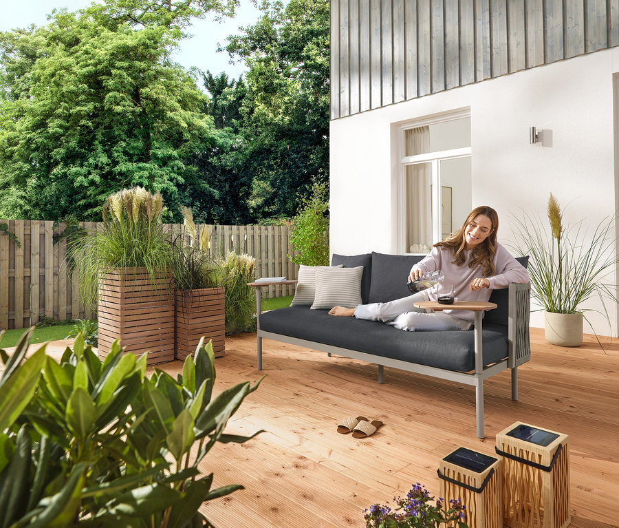 Žena nalévá vodu do sklenice na terase. Vedle ní stojí bambusová solární lucerna, malá a velká, solární zápichy s LED, 5 ks a zahradní pohovka se 2 otočnými stolky a truhlíky »Tinus«, 2 ks.