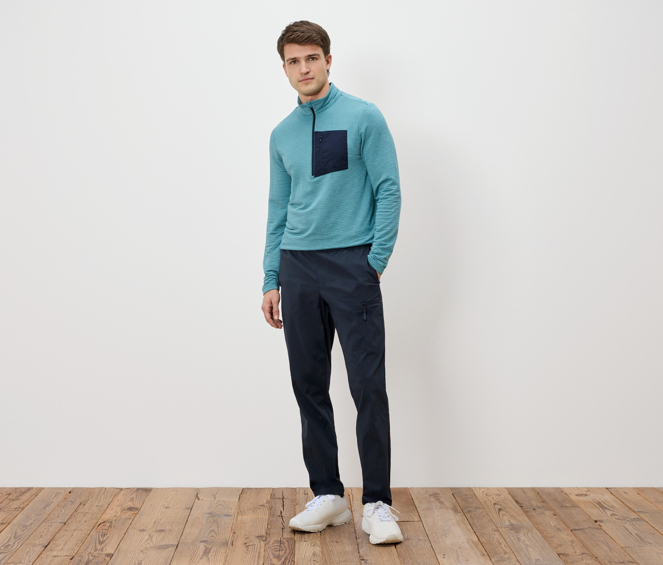 Muž stojí před bílou stěnou, má na sobě tyrkysové tričko s dlouhým rukávem s tmavou kapsou, tmavě modré kalhoty a bílé tenisky.