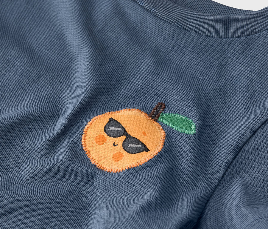 Detail modrého trička s našitou mandarinkou se slunečními brýlemi.