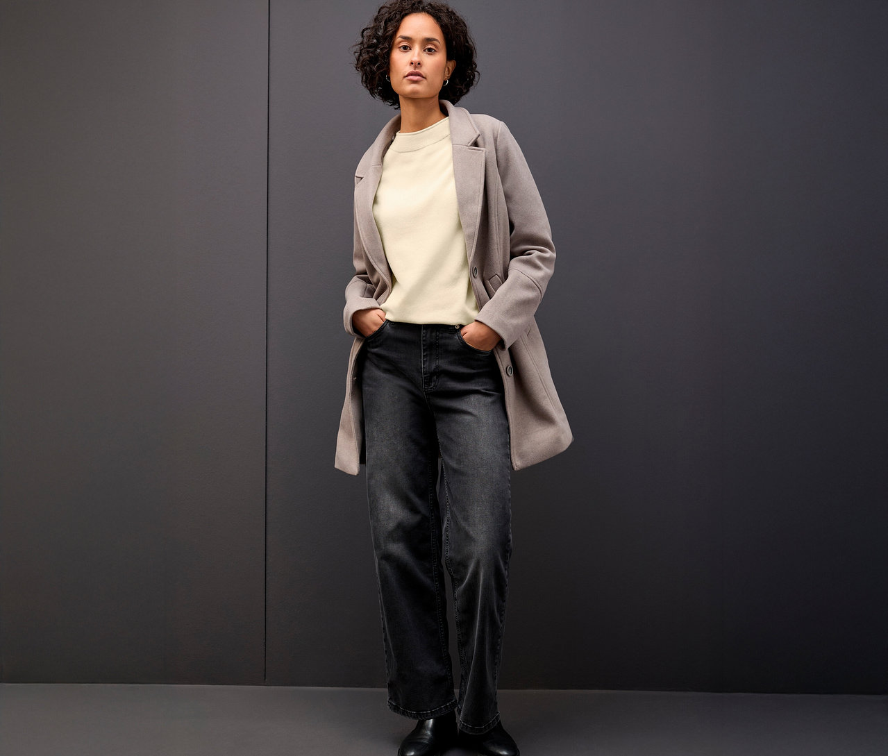 Žena stojí s rukama v kapsách a má na sobě béžový pletený svetr s rolovaným lemem Street One, šedou bundu a tmavé džíny.