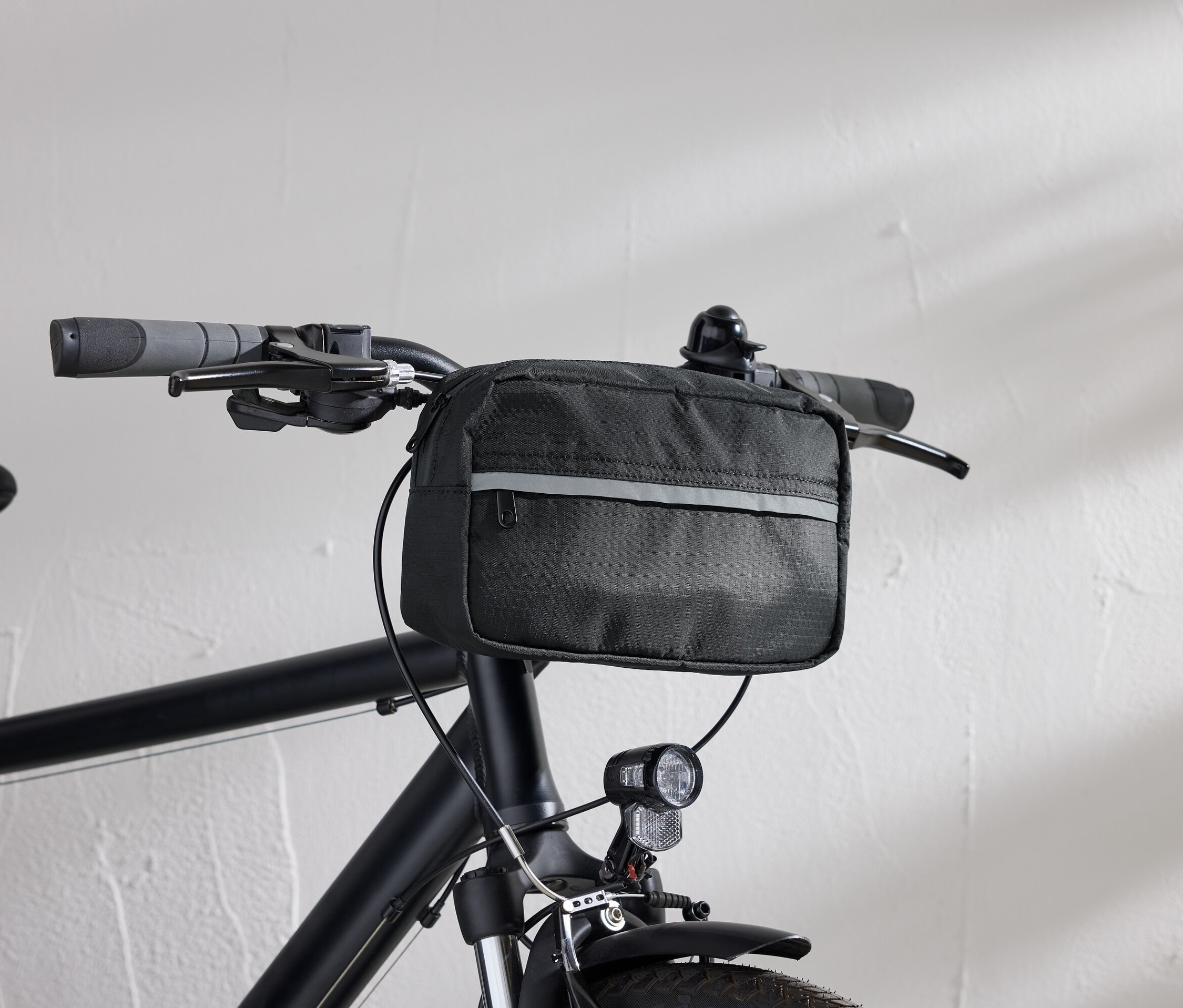Detailní pohled na černé kolo s černou brašnou na řídítka a světlem na kolo před bílou stěnou.
