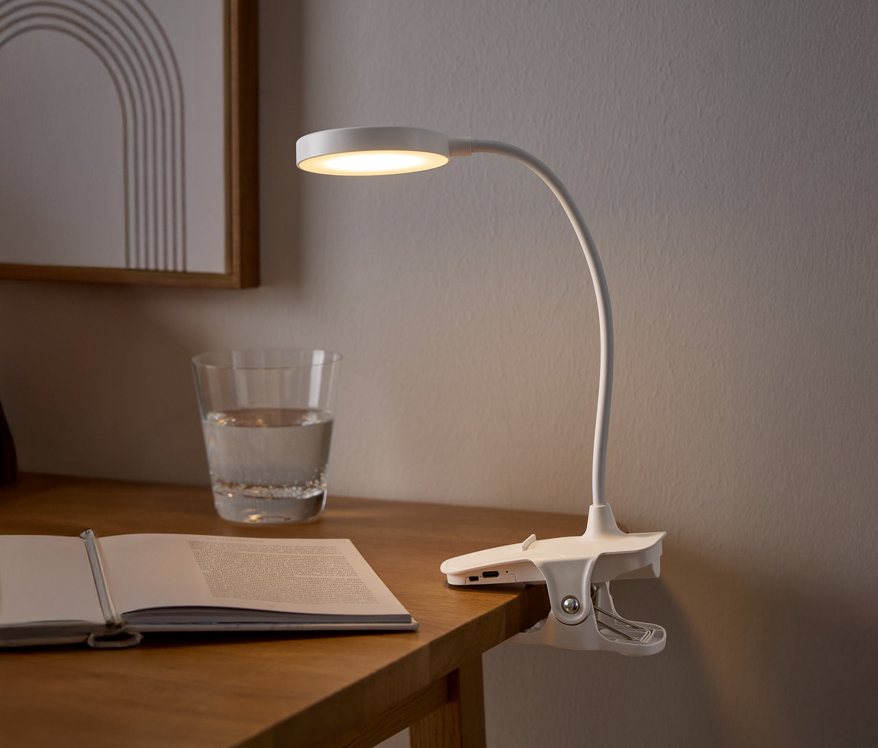 Bílá klipsová lampa osvětluje otevřenou knihu na dřevěném stole vedle sklenice vody a zarámovaného obrazu.