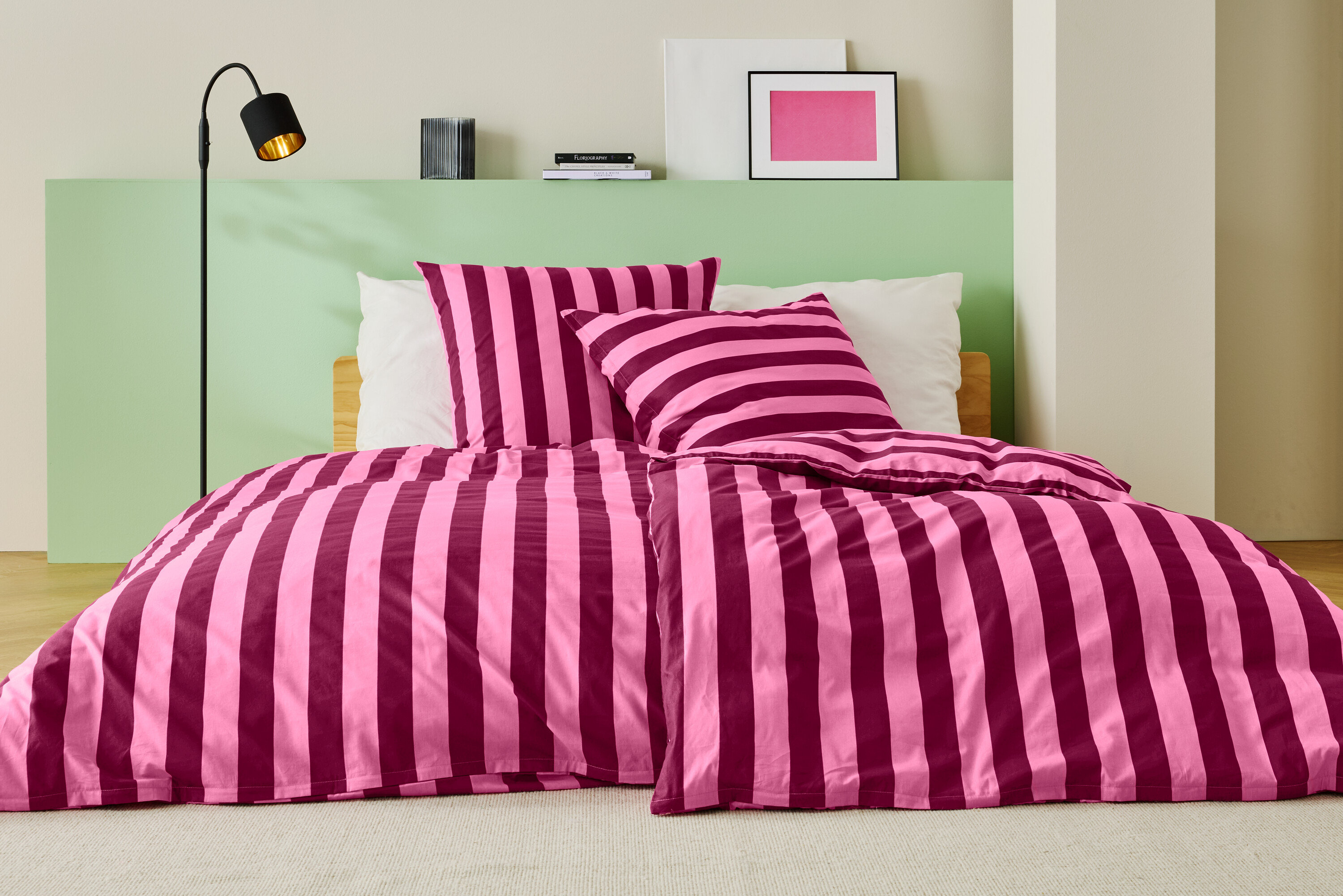 Postel s růžovo-vínově červeným pruhovaným ložním prádlem, dvěma polštáři a stojací lampou.