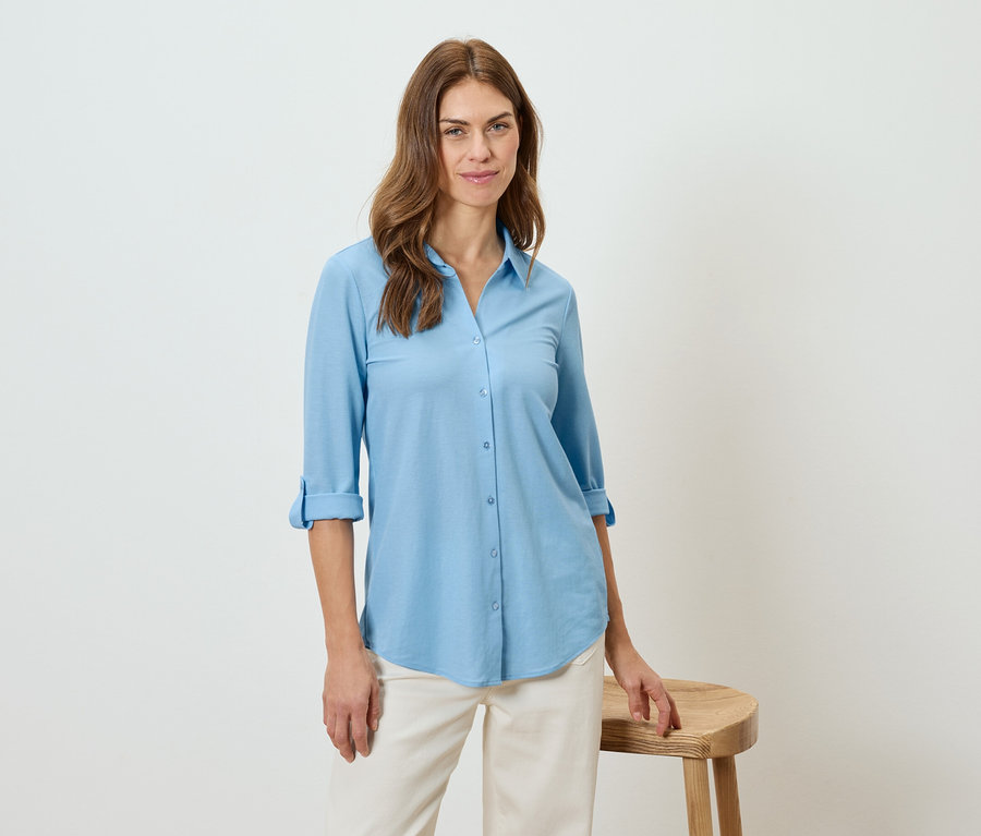 Žena má na sobě světle modrou košili a béžové kalhoty, opírá se o stoličku.