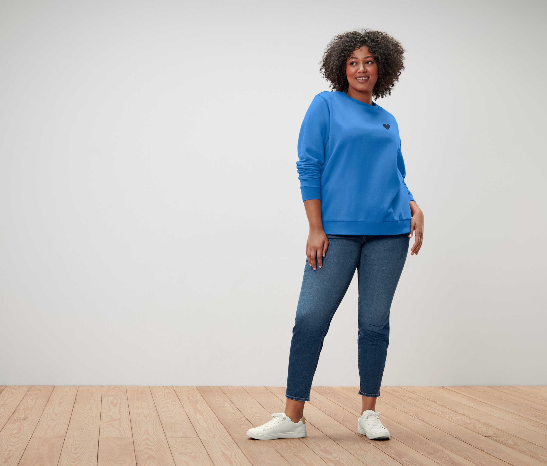 Žena s kudrnatými vlasy má na sobě modrou mikinu s malým černým srdcem, modré džíny a bílé tenisky.