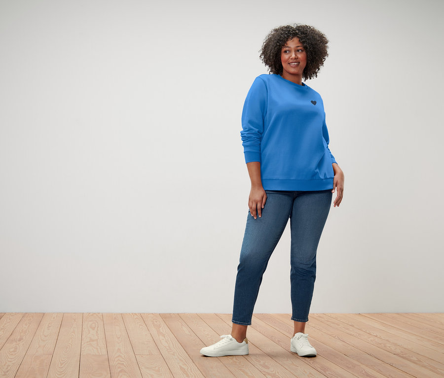 Žena s kudrnatými vlasy má na sobě modrou mikinu s malým černým srdcem, modré džíny a bílé tenisky.